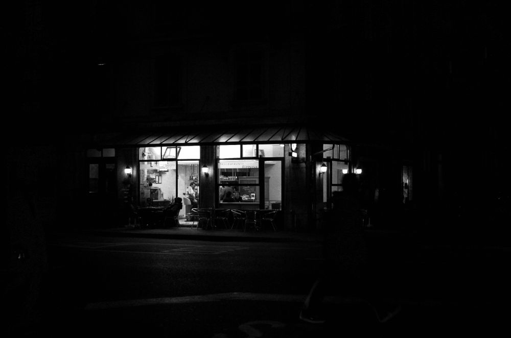 A b/w view of a pizzeria at night. The place is on a corner and all surroundings are dark except for the interior of the restaurant which is illuminated and with people inside.