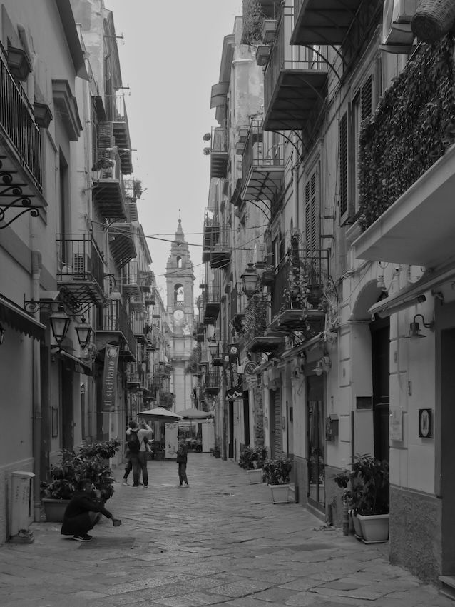 B/W image of a narrow street in central Palermo surrounded by buildings with balconies and plants. A few people in the middle of the pedestrian only street. A church steeple in the center background.