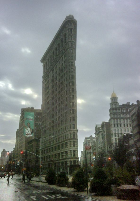 Flatiron building in Manhattan. Small trees on the pavement bottom right. Cloudy skies. 