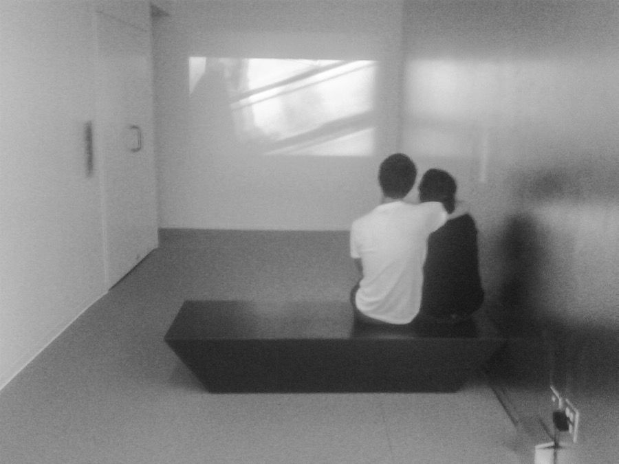 B/w image of a couple of viewers sitting together and watching a video on a narrow room.