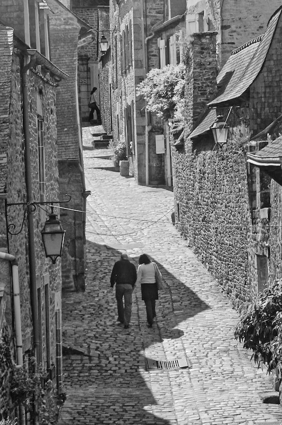 A b/w image of a cobblestone street with two persons walking. The narrow street surrounded by stone house. at the top center another person walking is also visible.