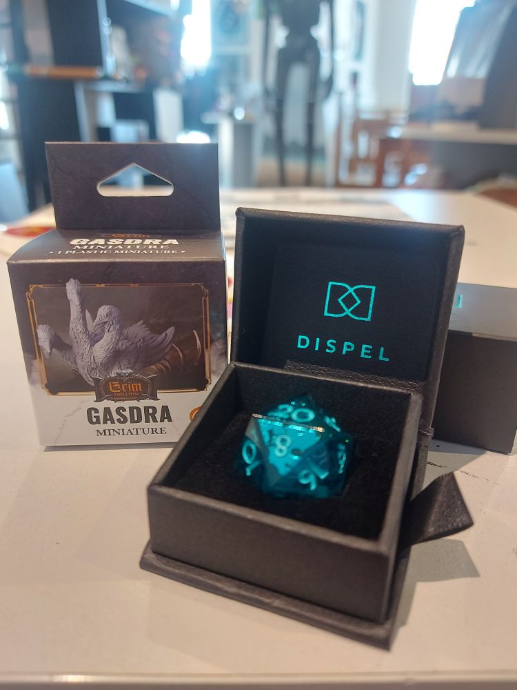 A boxed miniature of a 3 headed goose named Gasdra, along with a gorgeous glittery blue d20 sat in a black box. 