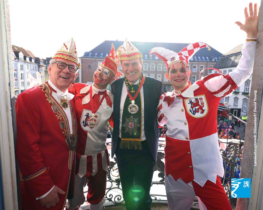Vier verkleidete Männer in bunten Karnevalskostümen posieren lachend auf einem Balkon vor einer Menschenmenge in einer Stadt.