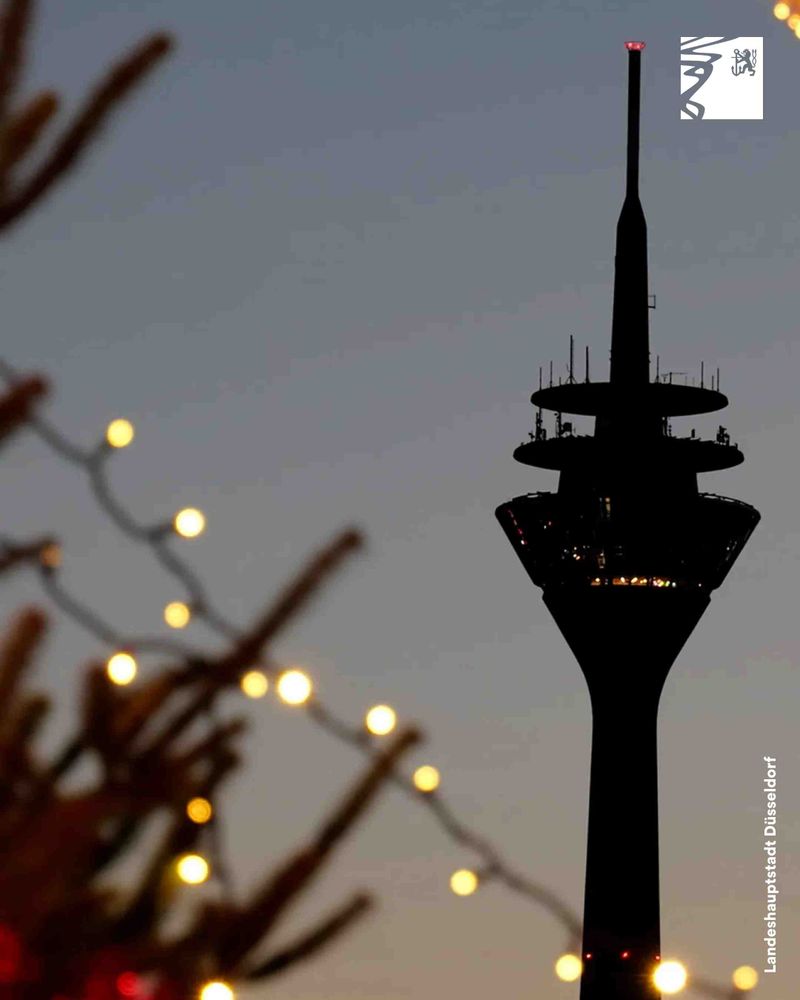 Silhouette des Rheinturms in Düsseldorf bei Dämmerung mit unscharfen Lichterketten im Vordergrund.