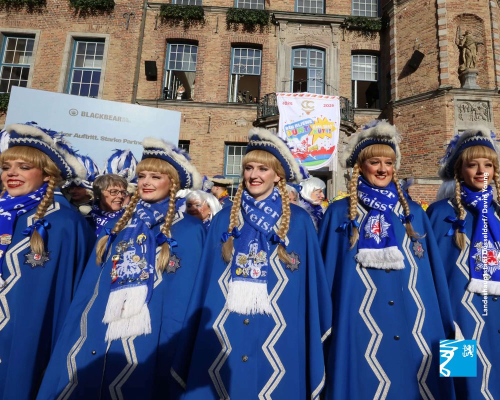 Gruppe von Menschen in blauen Karnevalskostümen mit weißen Verzierungen und Zöpfen vor einem historischen Backsteingebäude.