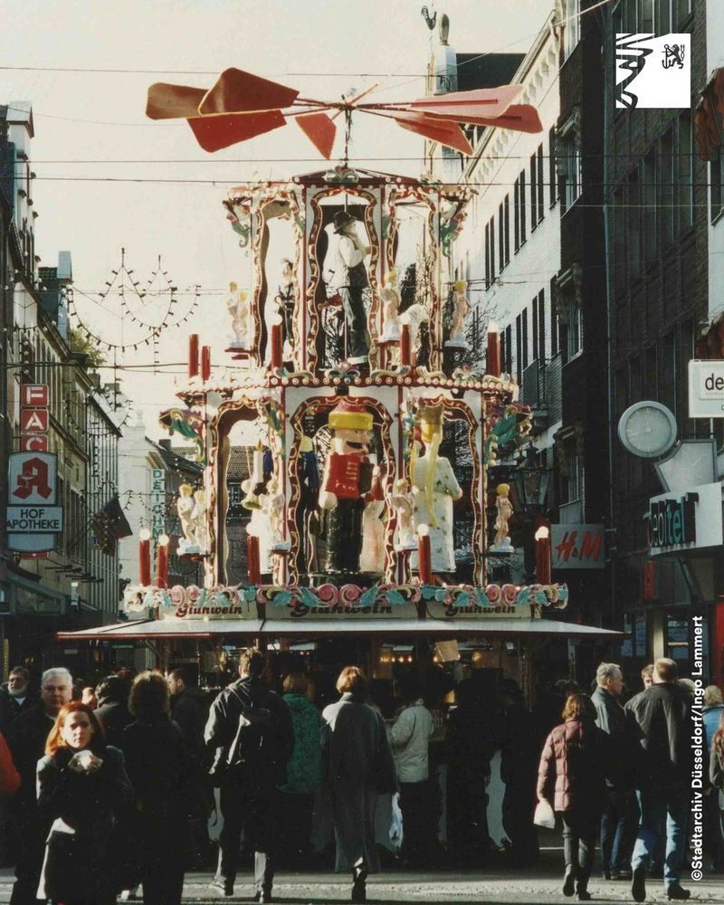 Großer, mehrstöckiger Weihnachtsmarktstand mit drehenden Figuren und rotem Propeller auf belebter Einkaufsstraße.