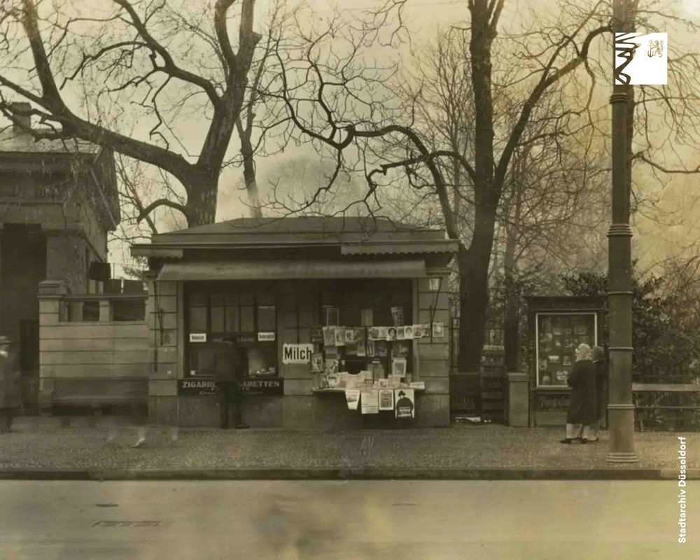 Altes Kiosk mit Milch- und Zigarettenverkauf vor kahlen Bäumen an einer ruhigen Straße im Winter.