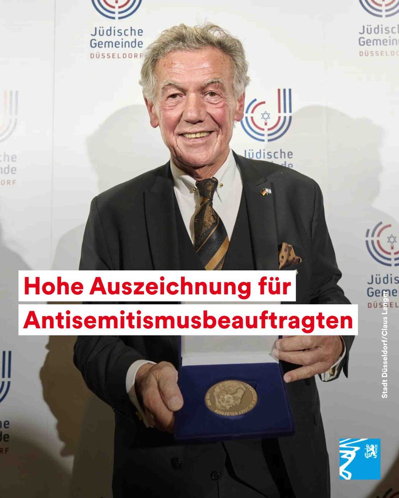 Mann in Anzug hält eine Auszeichnung vor einer Wand mit Logos der Jüdischen Gemeinde Düsseldorf.