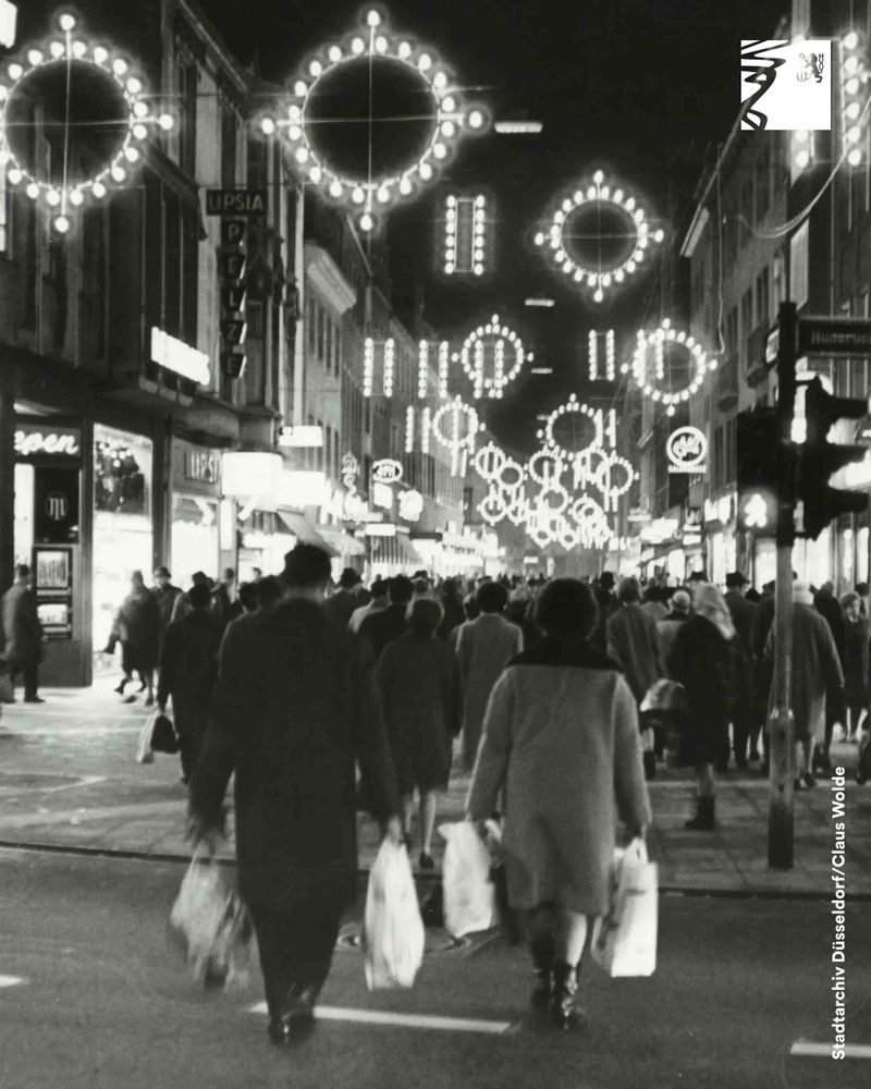 Menschen mit Einkaufstüten gehen abends durch eine beleuchtete Einkaufsstraße mit runden Lichterketten an der Decke.