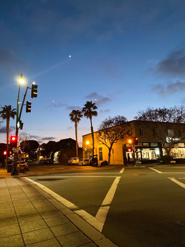 Waiting to cross the street during the evening. Palm trees around. The sky in blue and purple colors in the background. The moon up in the sky. Warm lights on the street. 