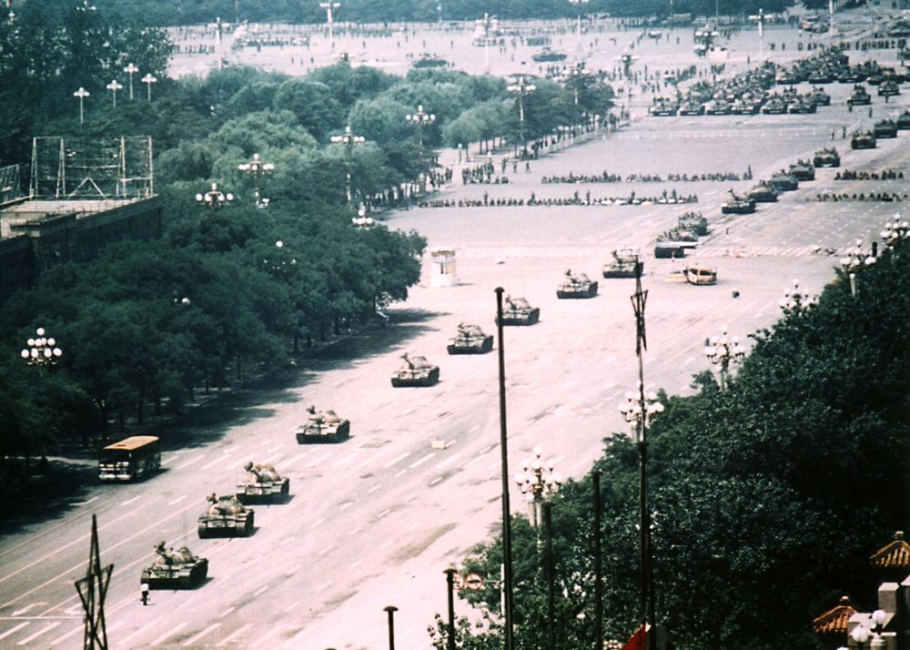 This wide-angle aerial photograph shows a broad avenue with a column of military tanks positioned down the center of the street. The tanks appear to be Type 59 or similar models, arranged in a single-file line stretching into the distance. In the foreground, a solitary figure can be seen standing directly in front of the lead tank, though from this elevated perspective the person appears quite small. The avenue is flanked by rows of trees on both sides, with large open plaza areas visible. In the background, there are multi-story buildings and what appears to be a large public square or gathering area. The photograph has the characteristic color tone and grain of film photography from the late 1980s.

This is the famous wide-shot perspective of the "Tank Man" incident at Tiananmen Square in Beijing, China, taken on June 4, 1989, capturing the moment when an unknown protester stood in front of a column of tanks following the government crackdown on pro-democracy demonstrations.

(ALT Text by Claude AI)
