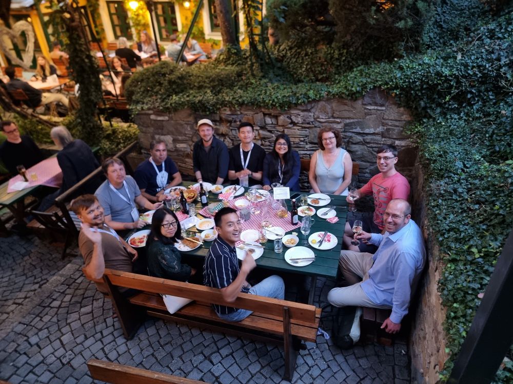 A group of twelve people sitting around a long outdoor dining table in a cozy courtyard restaurant with stone walls and greenery. They are enjoying food and drinks, smiling, and looking up toward the camera. Plates of food, wine glasses, and bottles are on the table, which is covered with a red-and-white checkered tablecloth. The atmosphere is warm and social, with other diners visible in the background.