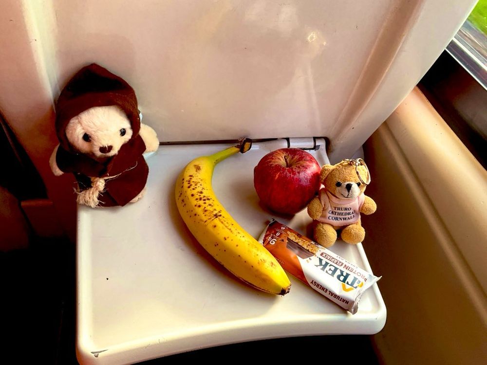 A monk bear in a brown habit, a banana, a red apple, a chocolate Trek bar, and Mary, on a fold down table on the back of a train seat