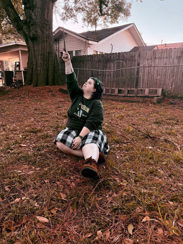 Me sitting in the grass with leaves and pine needles, I’m wearing a black white and grey plaid skirt and a long sleeve emerald green crewneck that says SLYTHERIN I’m wearing a emerald green Slytherin headband and I’m looking up my right arm is extended with a wand in it 