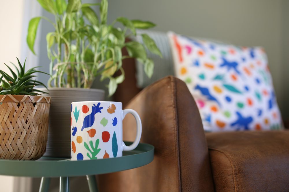 White ceramic mug with colourful abstract shapes on a green side table next to potted plants and a brown leather chair.