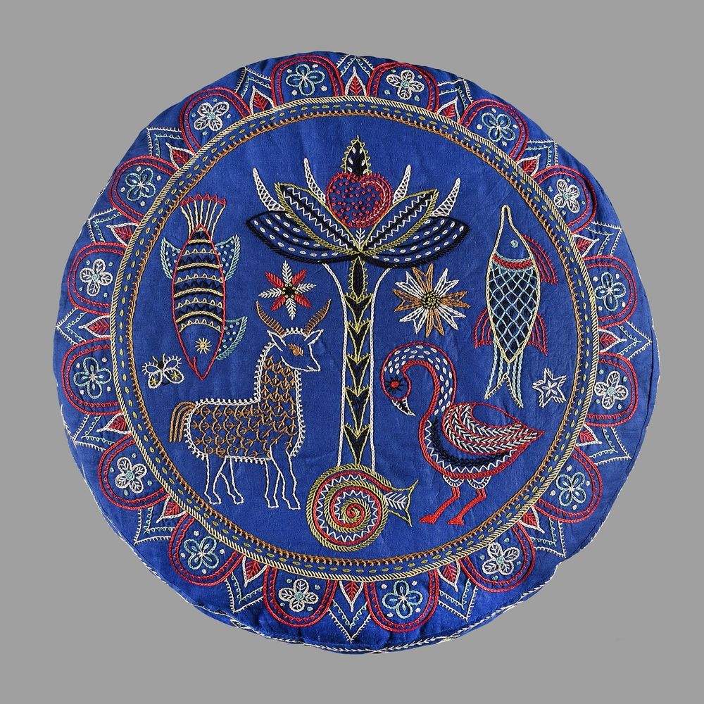 Round blue fabric with intricate embroidery of a deer, fish, bird, and floral motifs in red, white, and yellow thread.