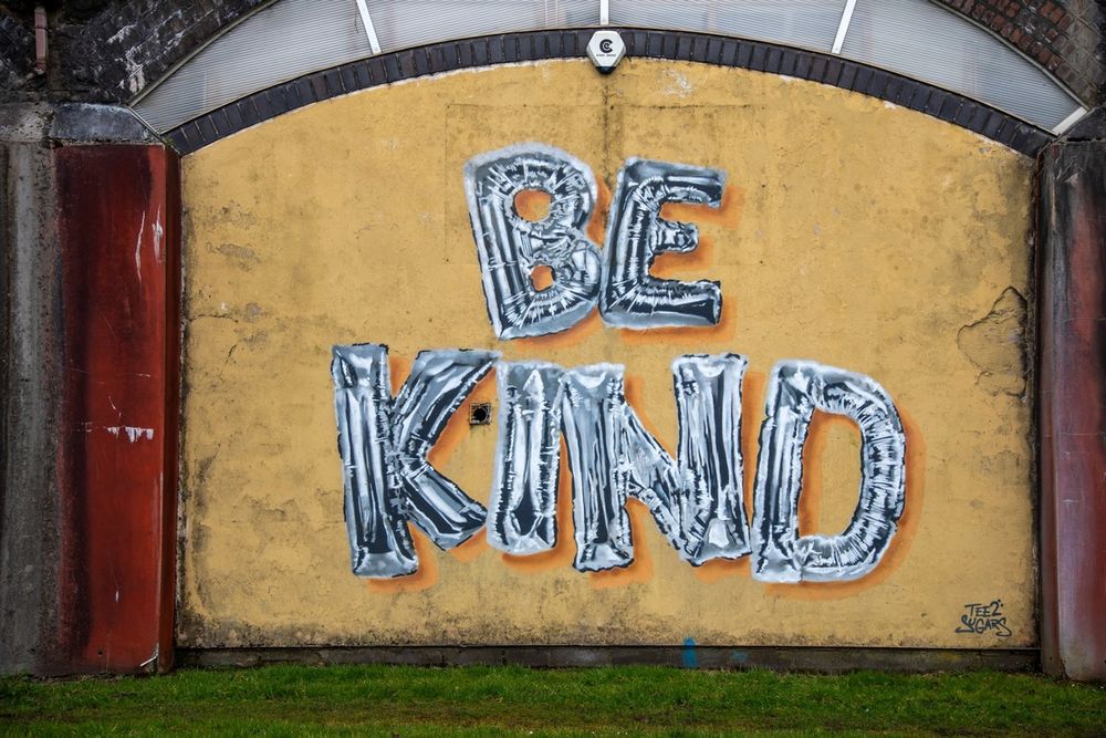 Mural on a yellow wall with the words 'BE KIND' painted in large silver balloon letters.