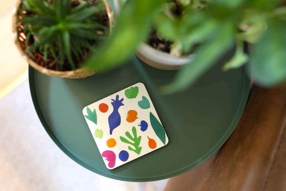 Colourful abstract shapes printed on a square coaster resting on a green round table by two potted plants.