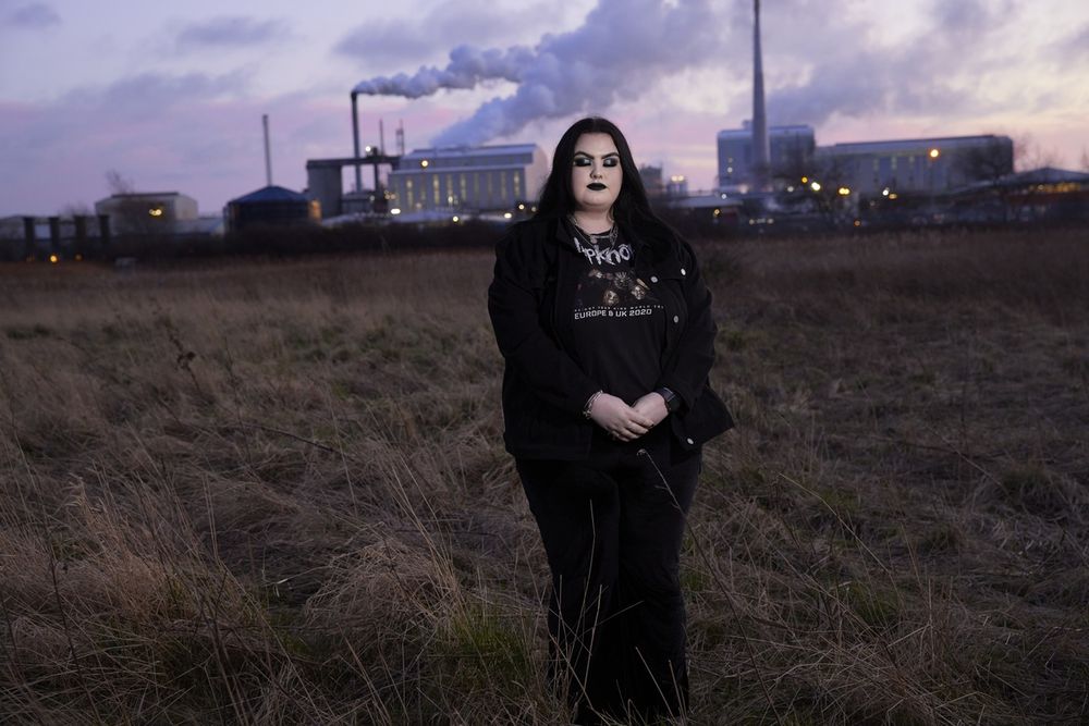 Person standing in a grassy field at dusk with an industrial plant emitting smoke in the background under a cloudy sky.