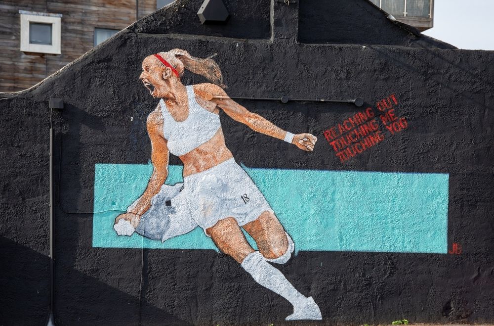 Mural of a Chloe Kelly in white uniform mid-action against a black wall with a turquoise stripe and red text.