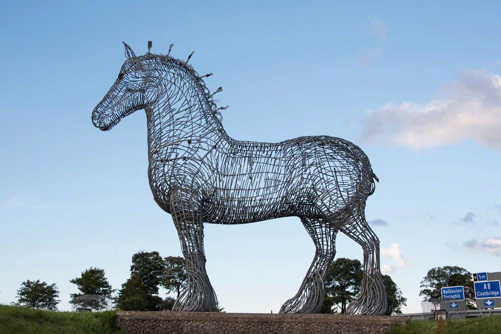 Large steel frame sculpture of a horse standing on a grassy hill under a clear blue sky.