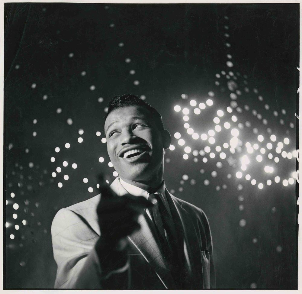 A black and white photograph of a black man dressed in a suit with a big grin on his mouth. The camera angles up to him with fairy lights filling the background.