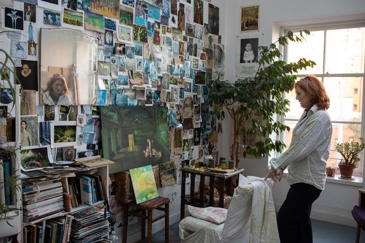 Artist's studio filled with numerous pinned photographs and paintings covering the walls, with a woman standing near a window beside a large leafy plant.