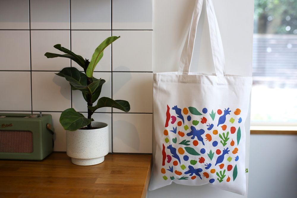 White tote bag with colourful abstract shapes hanging on a white wall next to a potted green plant on a wooden countertop.