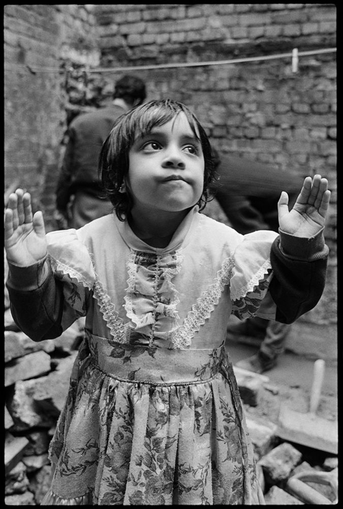Child in a floral dress with lace details raising both hands in a rustic brick courtyard.