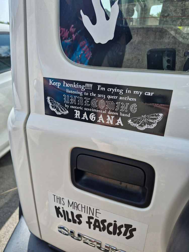 A white Suzuki jimny rear door with stickers on it. 

The top sticker reads "Keep honking!!! I'm crying in my car listening to the queer anthem Unbecoming by esoteric sentimental doom band Ragana"

The bottom sticker reads "This machine kills fascists"