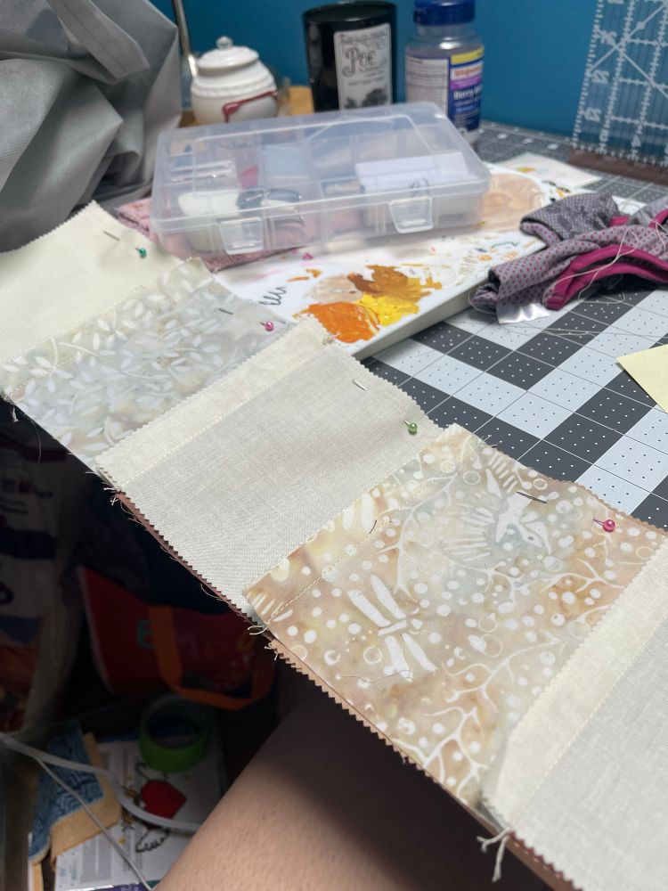 The original row for the quilt which features an off white cotton or line. With a lightly dyed and patterned wash, pins are holding it with another row before it is sewn. 