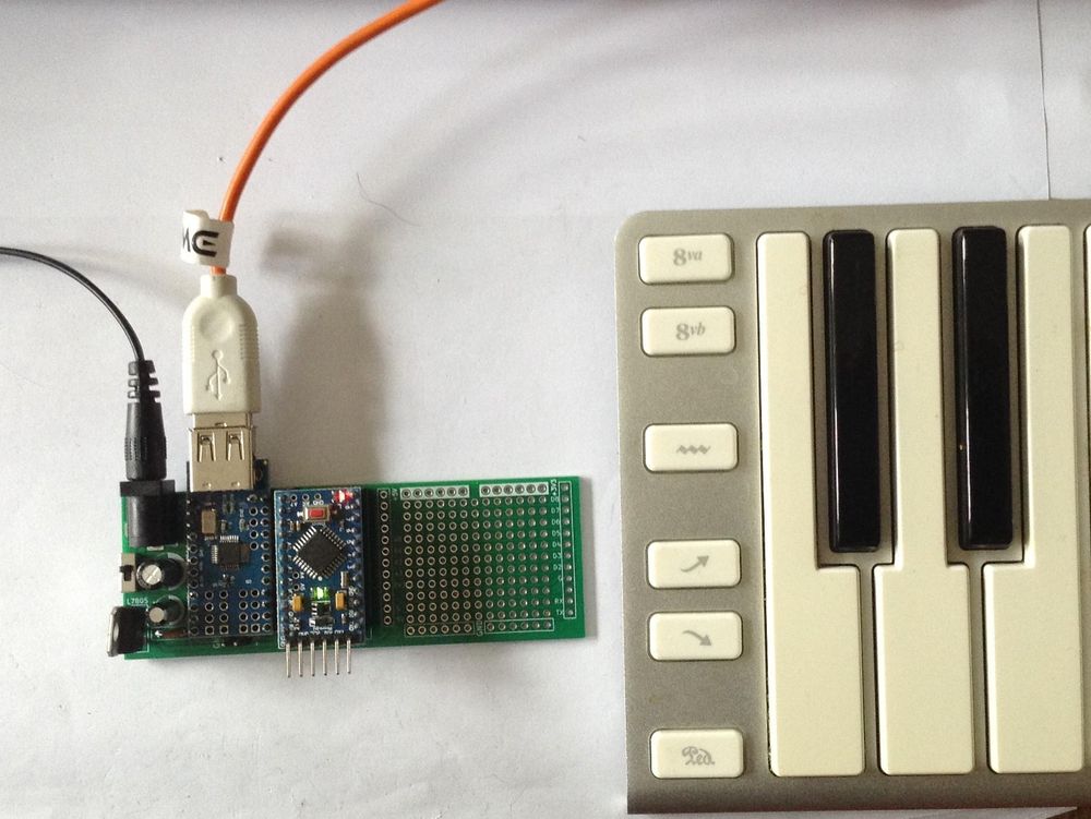 Photo showing the described DIY PCB in use.  It is a rectangular PCB 100x35 mm.  An Arduino Pro Mini and mini USB Host shield are mounted on the board.  The board has power from a barrel jack and the photo shows a USB MIDI keyboard plugged into the USB socket on the mini USB Host shield.