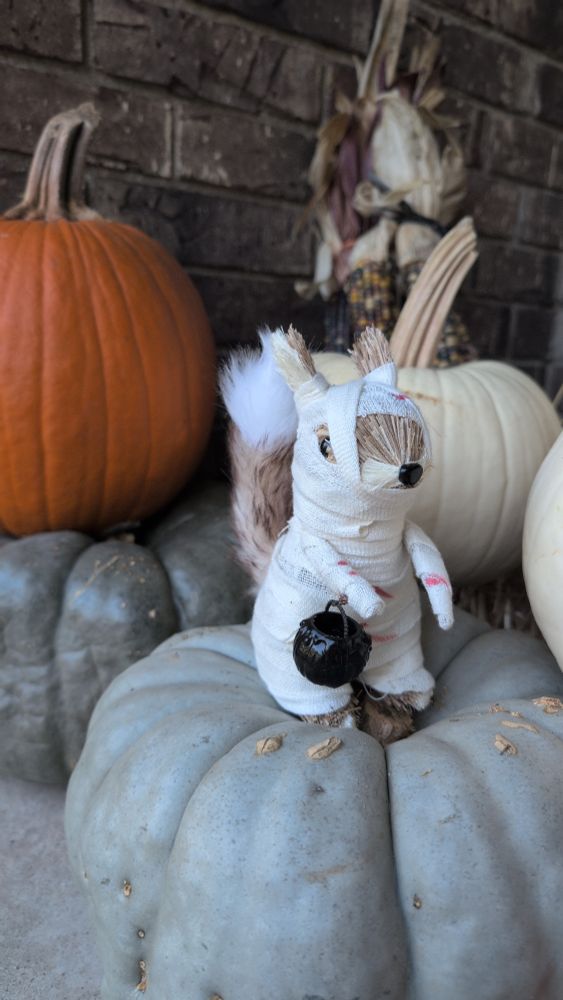 A Halloween decoration that is a squirrel dressed as a mummy and carrying a pumpkin trick or treat bucket. 