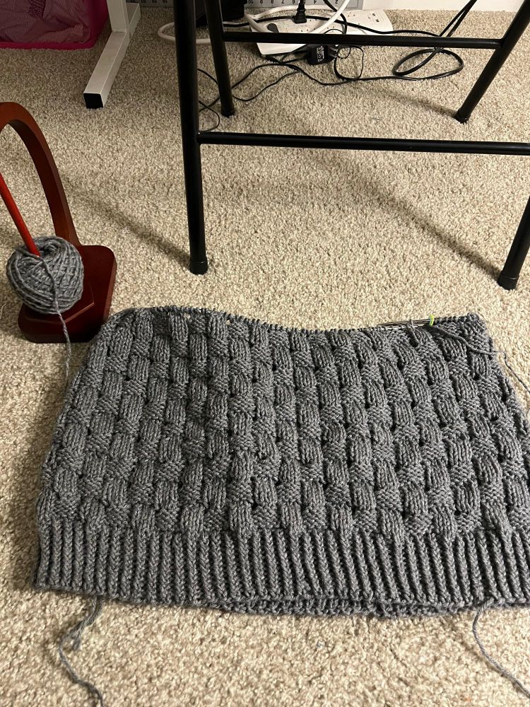 Bottom part of the Korshavn sweater in grey linked to a yarn stand with a ball of grey yarn on it 