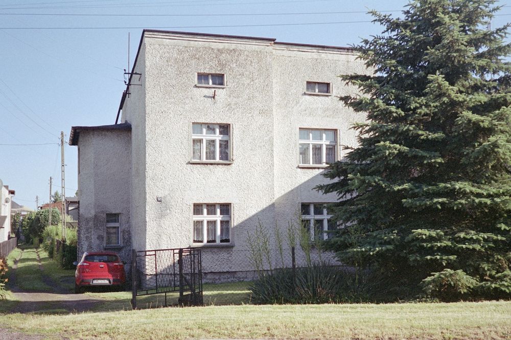 a large square house partially obscured by a large evergreen on one side, and a red car parked on the other