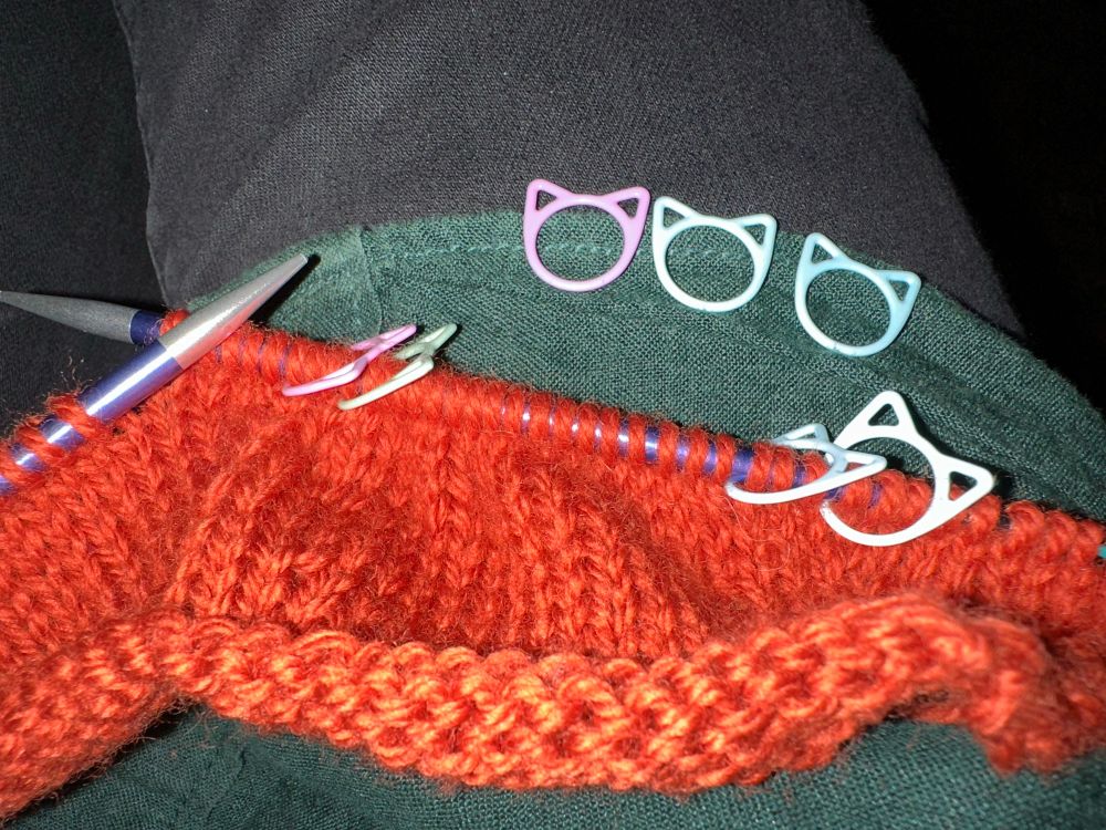 A photo of an orange knitting project (it’s a cardigan) still on the needles with cute little stitch markers shaped like cats’s heads. 