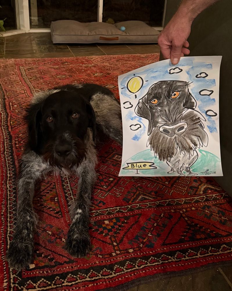 A handsome dog sits on the carpet beside a drawing of himself. 