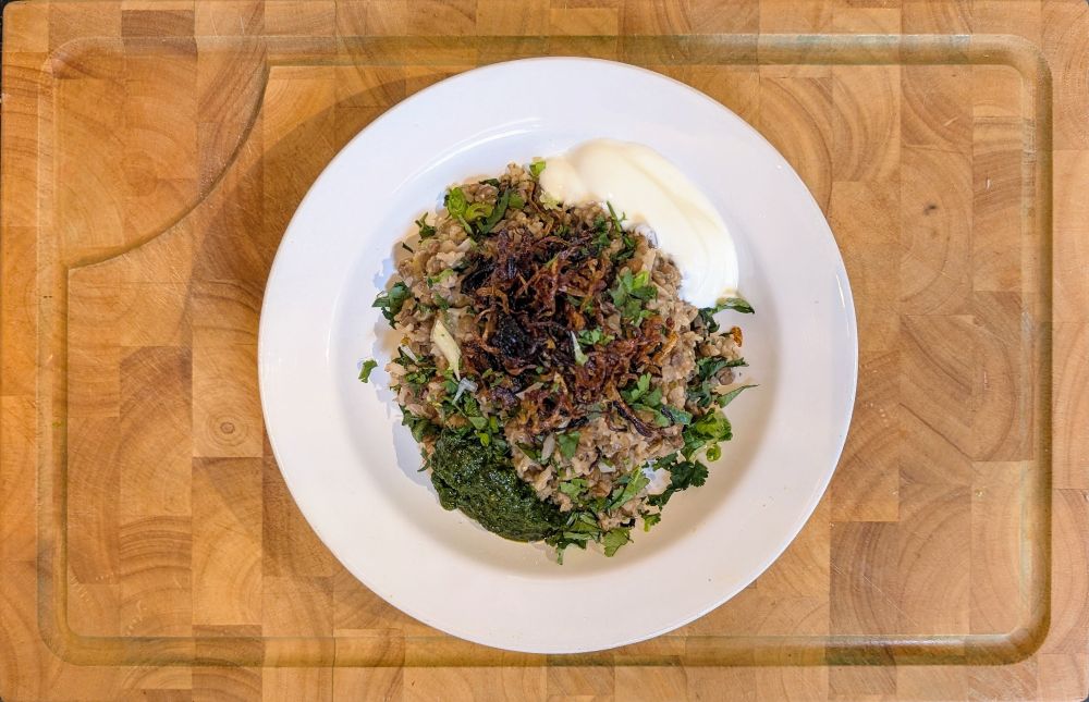 Photo of a dish of Mujaddara, a mound of brown basmati rice and lentils, topped with richly caramelised, crispy fried onions, chopped scallions and cilantro. On either side of the pile of rice and lentils is a sauce - zhoug, a dark green coriander and chilli sauce on one side, a generous spoonful of Greek-style yoghurt on the other.