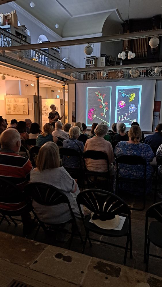 Lady giving a talk to an audience who are seated in a darkened museum. Large images of flowers on a black background. 