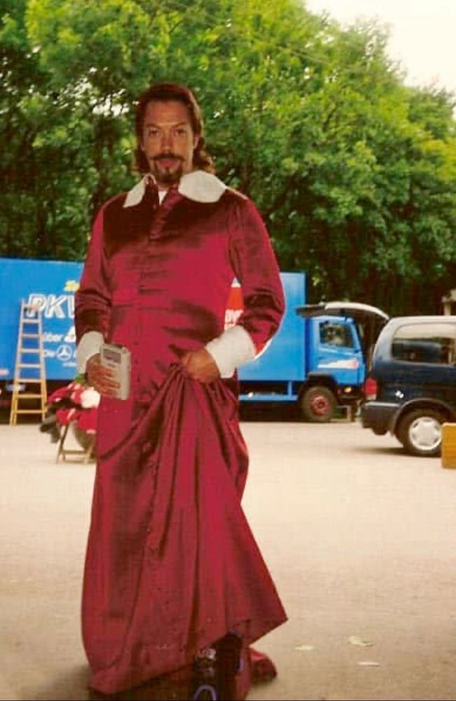 tim curry dressed up in a swaggy medieval-looking red robe holding a game boy. a truck, a car and a stepladder are visible in the background. he kind of looks photoshopped in but it's probably just his raw tim curry energy