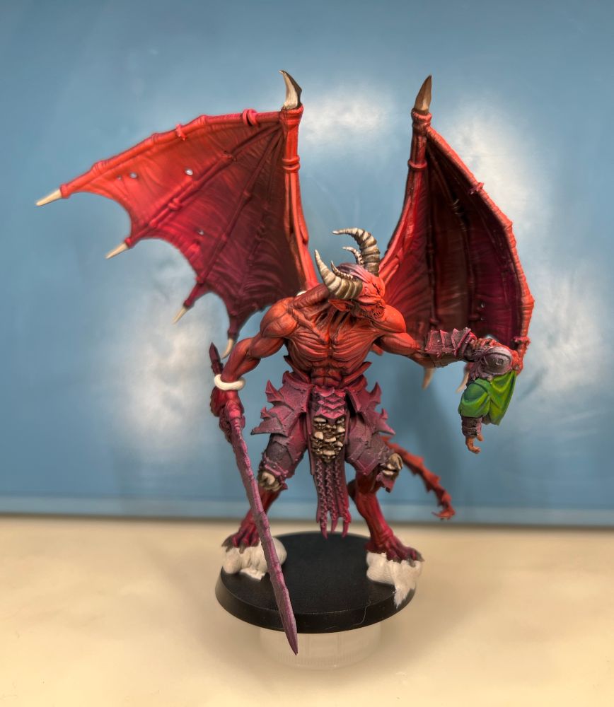 A red demonic humanoid is holding the upper half of a dwarf’s body and staring into his eyes. Its body is partially painted and is red. The dwarf is largely unpainted except for its green cloak. Designed by Den of Imagination. Front view. 