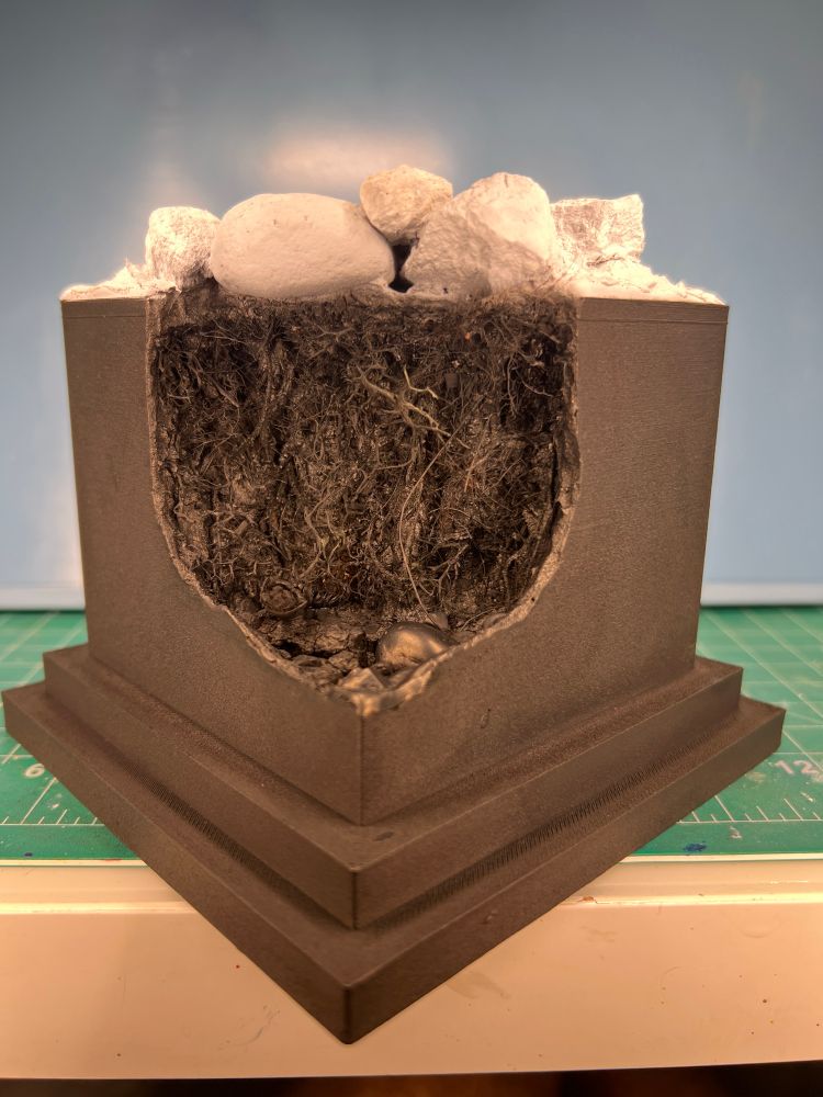 Black plinth/base with the corner cut out and filled with miniature vegetation, pebbles, and rocks, all painted black. The top is now painted white with the miniature boulders prominent.