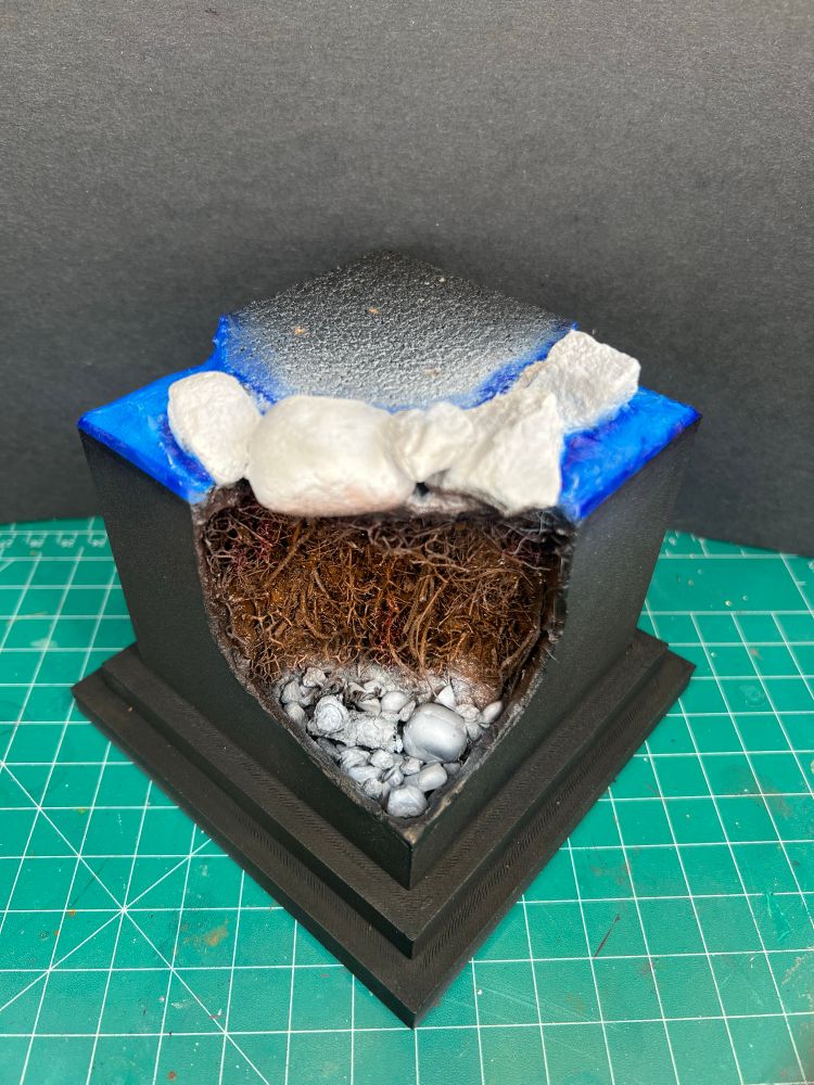 Black plinth/base with the corner cut out and the vegetation is painted brown, and the pebbles, small rocks, and logs are now painted in a gradient of grey and white. On top, the miniature boulders are painted grey and white, and the open area is painted in light and darker blue. The brown is Kimera Kolors oxide brown light, the blue is Daler Rowney Inks Rowney Blue and Golden High Flow Acrylics indigo. 