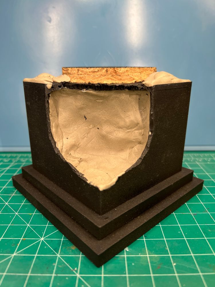 Black plinth/base with the corner cut out and beige clay-like material filling it. Flat cork board visible on top, with raised areas made of the same clay-like material. That material is Apoxie Sculpt. 