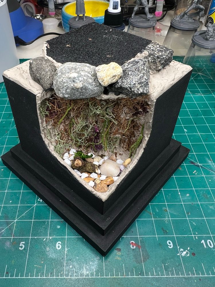 Black plinth/base with the corner cut out and filled with vegetation and pebbles with small rocks. There are two miniature logs visible. On top are five miniature boulders. The flat cork on top is now painted black. 