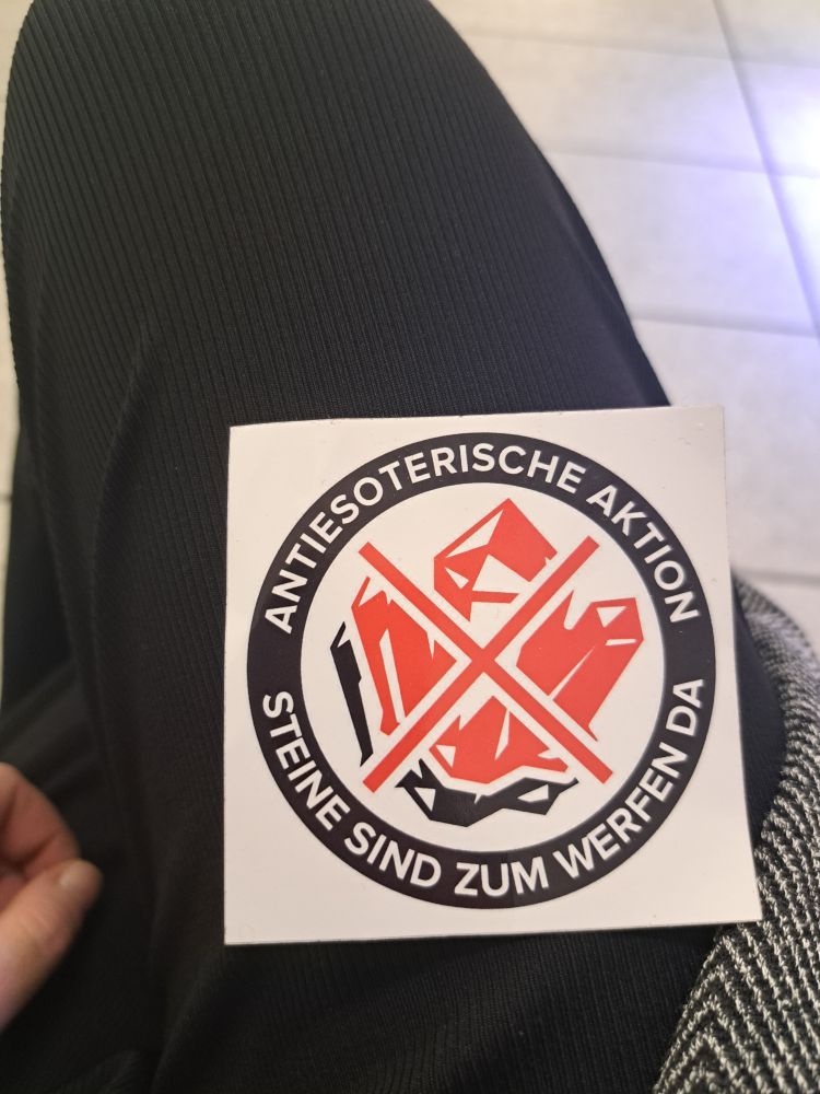 Das Bild zeigt einen schwarz roten sticker auf weißem Grund.
Ein schwarzer Kreis mit weißen Schriftzug "antiesoterische Aktion, Steine sind zum werfen da" umrandet ein Bild von roten durchgestrichen Kristallen. 