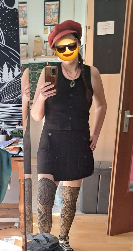 Eine weibliche Person macht ein Ganzkörperselfie vor einem Spiegel.
Sie trägt eine schwarze Kombi aus Rock und kurzer ärmelloser weste und eine orangene Kappe.
Das Gesicht ist mit einem Sonnenbrille-Emoji unkenntlich gemacht. 