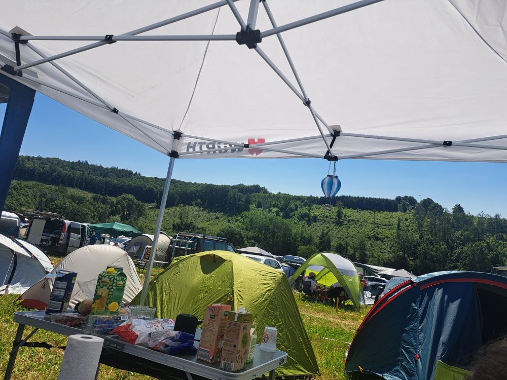 Blick auf einen Festival-Campingplatz. Im Hintergrund sieht man einen Wald und strahlend blauen Himmel. 