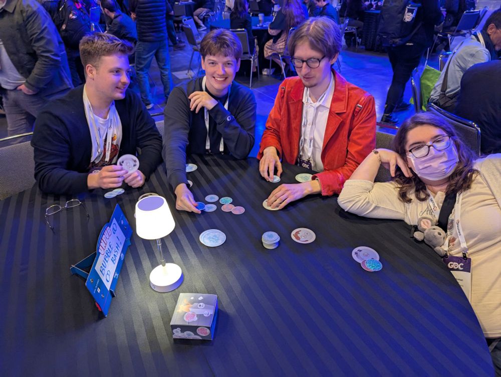 Four people sitting at a table playing a card game with various chips. A small lamp and a box are on the table. One person is wearing a mask. The setting appears to be a busy event.