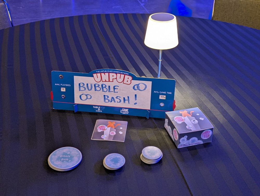 Table with "Unpub, Bubble Bash" game display, including a sign, circular game cards, and a small box, under a small white lamp on a striped tablecloth.